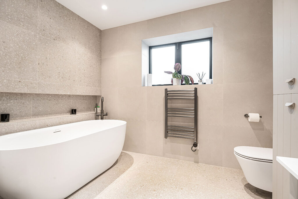 Modern bathroom with a freestanding white bathtub, wall-mounted towel warmer, toilet, and a large window with potted plants; neutral-colored walls and floor create a clean, minimalist look.