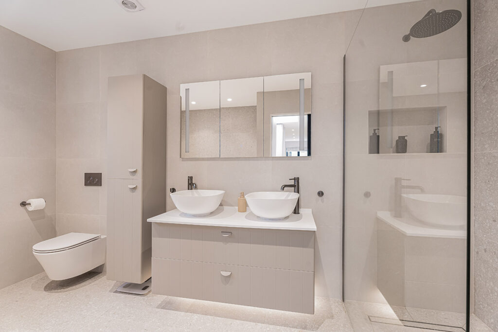Modern bathroom with beige walls, a wall-mounted toilet, a tall cabinet, a double-sink vanity with vessel sinks, large mirrored cabinets, and a walk-in glass shower with a rainfall showerhead.