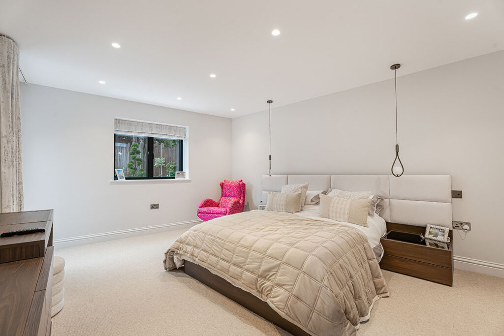 Modern bedroom with a large bed, neutral bedding, bedside tables, hanging pendant lights, a small window, and a bright pink chair in the corner. The room has beige carpet and white walls.