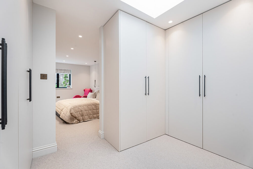A modern, minimalist bedroom with white fitted wardrobes, beige carpet, and recessed lighting. The bed has a beige quilt and bright pink cushions, and a window with greenery outside is visible in the background.