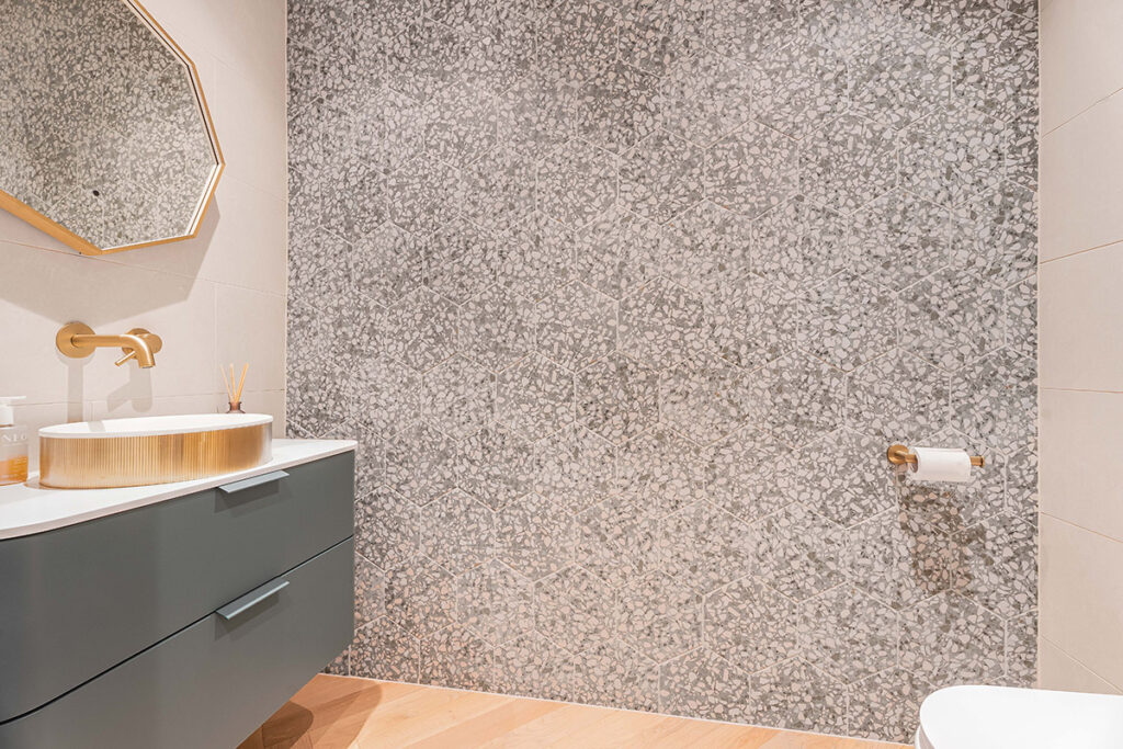 Modern bathroom with a geometric mirror above a sink with a gold faucet and round basin. The wall behind features a gray mosaic tile design, and a toilet paper holder is mounted on the right wall.