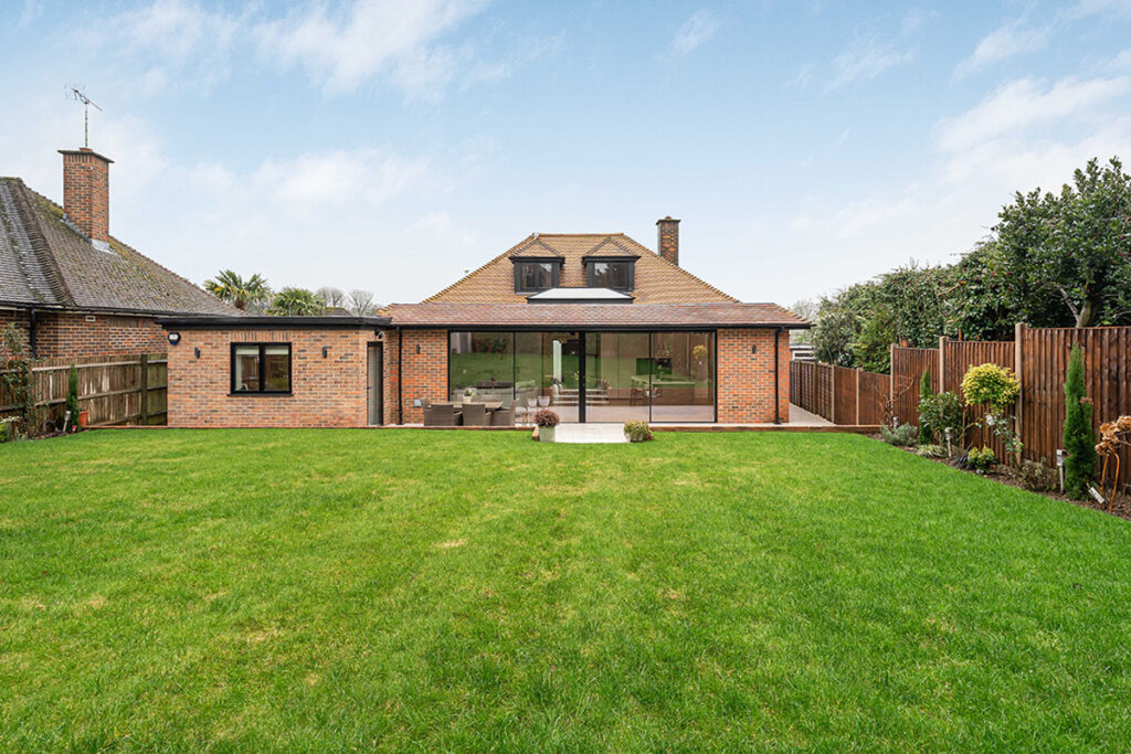 A modern brick house with large glass sliding doors opening to a spacious, well-kept green lawn, bordered by wooden fences and a small garden, under a partly cloudy sky.
