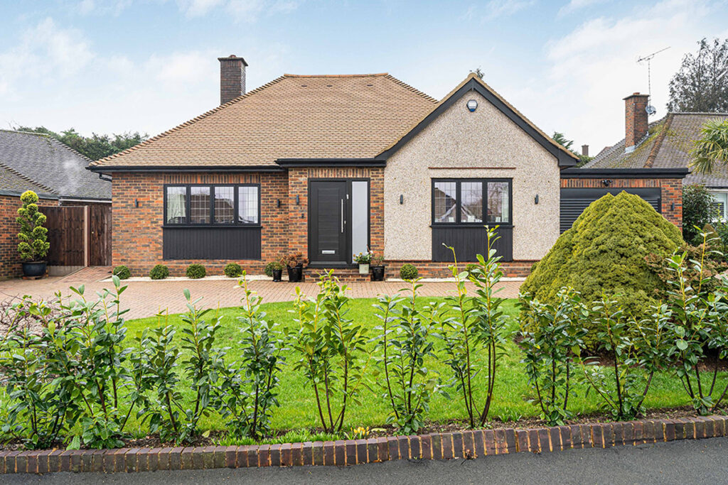 A single-story brick house with a beige tiled roof, black-framed windows and doors, and a tidy front lawn bordered by shrubs and a low brick wall. A paved driveway leads to a wooden gate on the left.