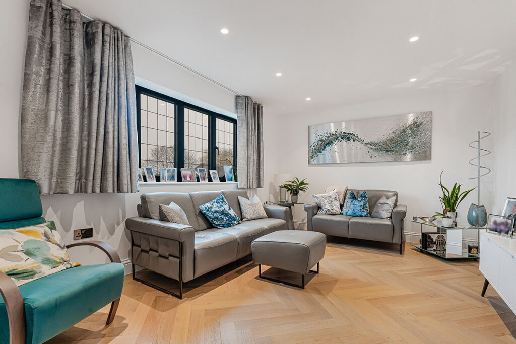 Modern living room with wooden herringbone floor, grey sofas, teal armchair, and large window. Decorative pillows, plants, and abstract wall art add color. Natural light brightens the clean, stylish space.
