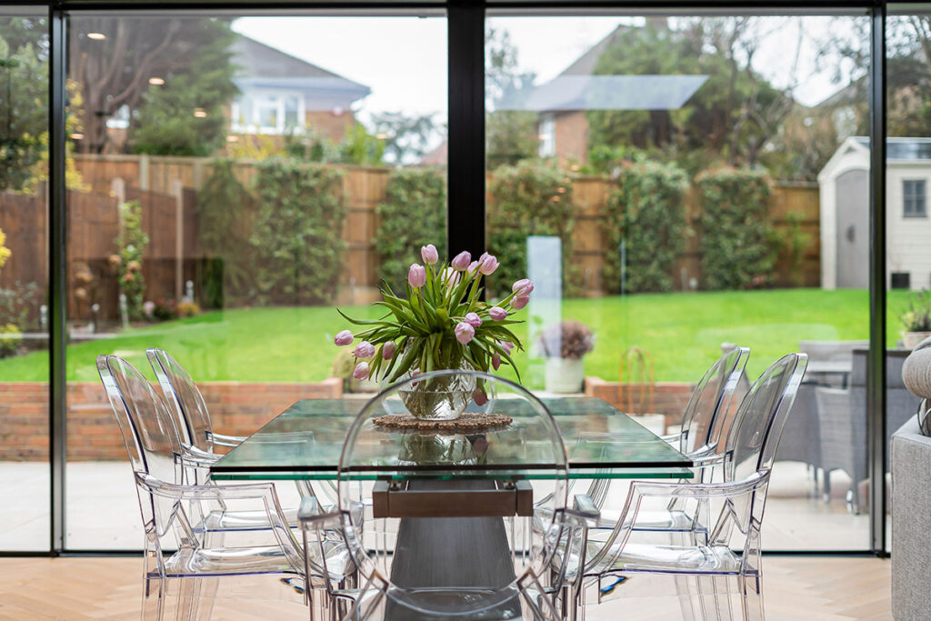 A glass dining table with a vase of pink tulips is surrounded by six transparent chairs, set in front of large windows overlooking a green backyard garden.