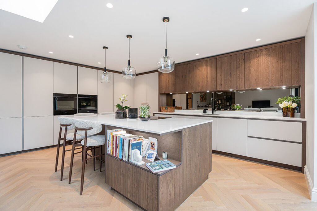 Modern kitchen with a large island featuring books and plants, bar stools, pendant lights, wooden cabinets, built-in oven, and light herringbone wood floors. The space is bright and contemporary.