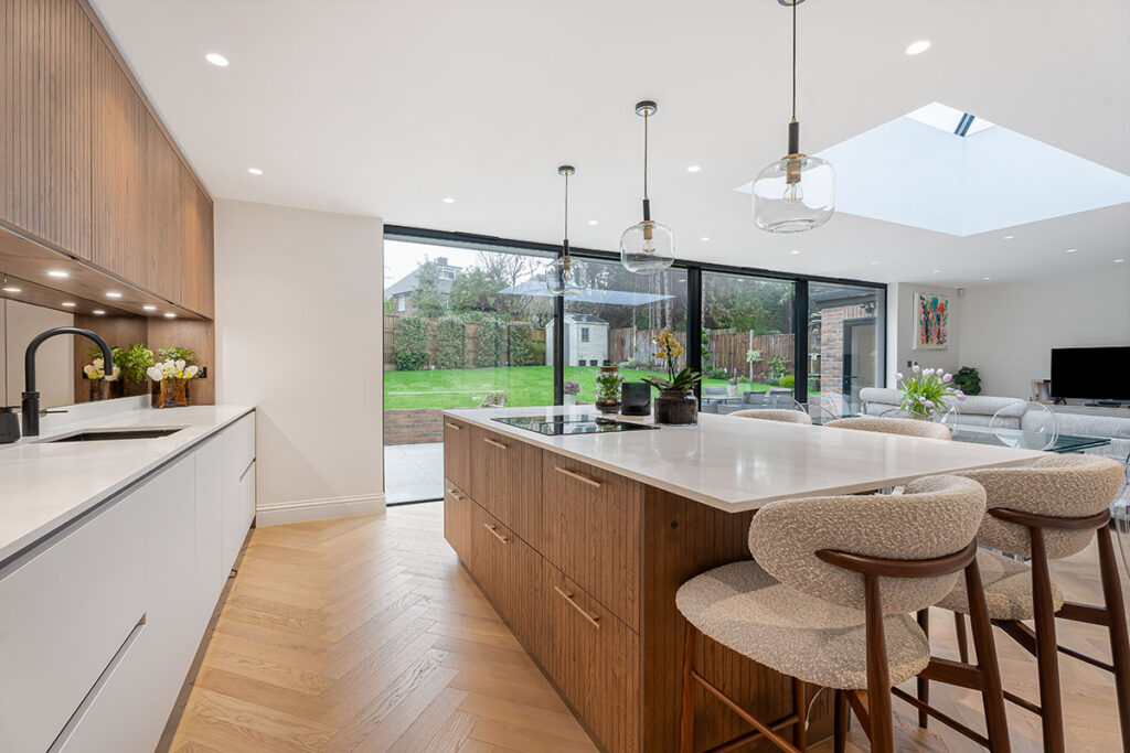 Modern open-plan kitchen with wood and white cabinetry, a large island with bar stools, pendant lights, and floor-to-ceiling glass doors overlooking a garden. Living and dining areas are visible in the background.