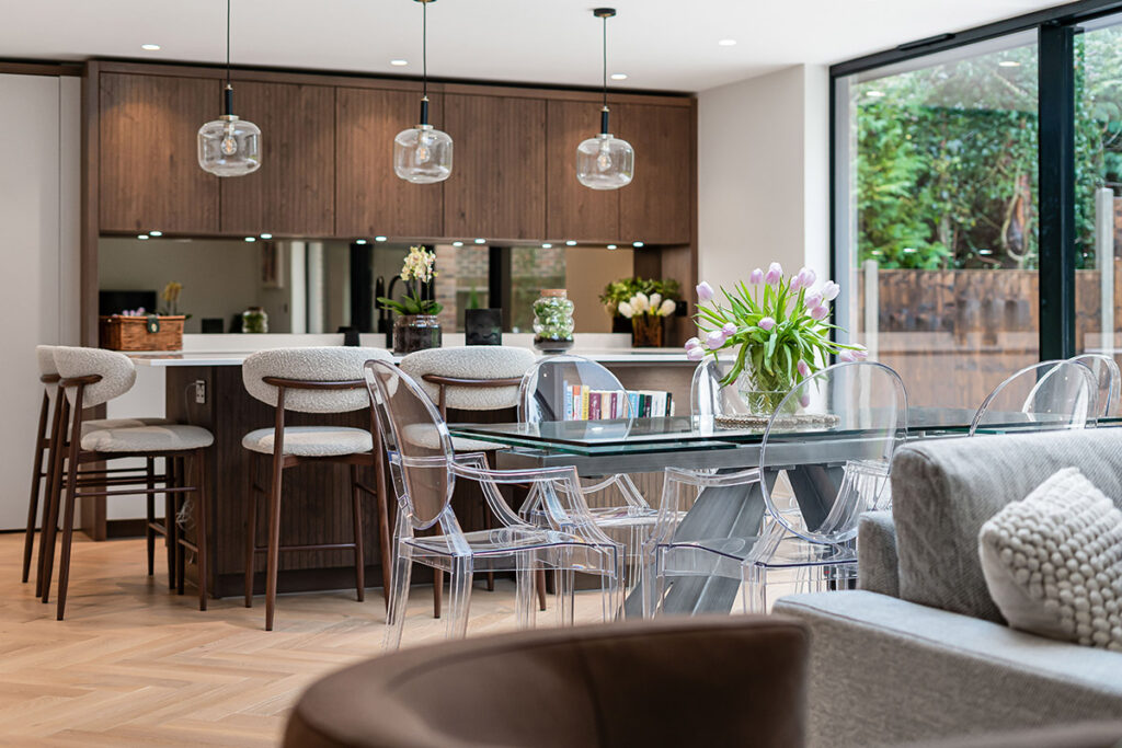 Modern open-plan kitchen and dining area with wooden cabinets, an island with bar stools, glass dining table with transparent chairs, fresh flowers, pendant lights, and large windows overlooking a garden.