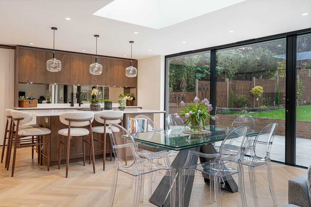 Modern kitchen and dining area with wood cabinets, a white island with bar stools, glass pendant lights, a glass dining table with clear chairs, and large sliding doors opening to a garden patio.