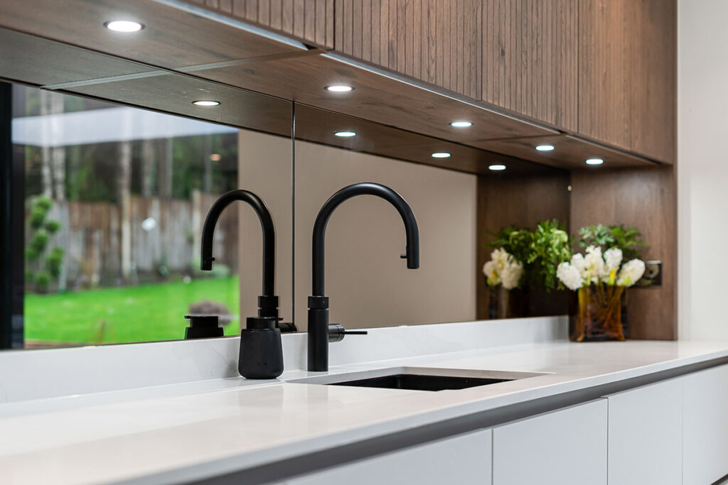 Modern kitchen with a white countertop, black faucet, soap dispenser, and mirrored backsplash. Wooden cabinets above are lit by recessed lights. Vases with white flowers are visible beside the sink.