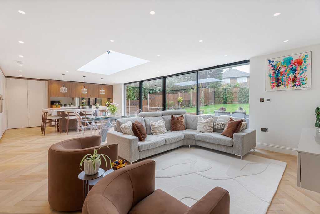 A spacious, modern open-plan living area with a large gray sectional sofa, two brown armchairs, a white rug, and floor-to-ceiling windows overlooking a backyard garden. A dining area and kitchen are visible in the background.
