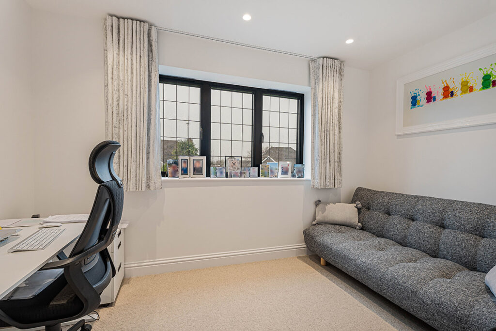 A bright home office with a desk, computer, and ergonomic chair on the left, a gray tufted sofa on the right, and family photos on the windowsill beneath large, black-framed windows. Colorful art hangs on the wall.