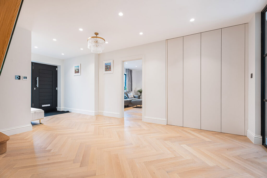 Modern, spacious hallway with light herringbone wood flooring, white walls, recessed lighting, a black front door, built-in closets, wall art, and a crystal chandelier; an open doorway leads to a bright living room.