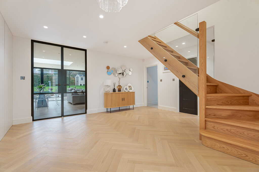 Spacious modern hallway with wooden herringbone floors, a wooden staircase with glass railing, a sideboard with decor, and glass doors leading to a bright living area and garden.
