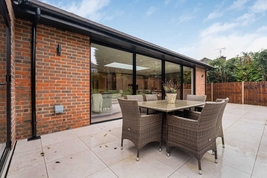 A modern patio with wicker dining set, cream tiles, and floor-to-ceiling windows reflecting a bright sky, attached to a red-brick building and surrounded by a wooden fence and greenery.