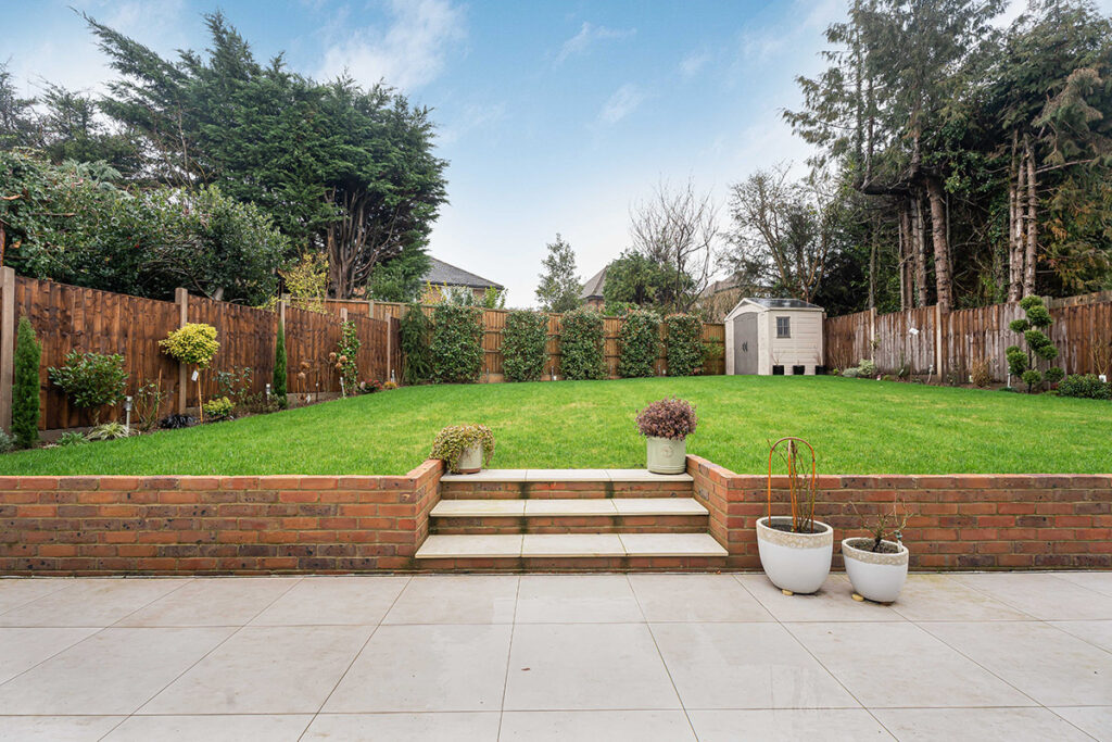 A neatly landscaped backyard with a green lawn, wooden fence, small shed, and potted plants on a tiled patio with steps leading to the grass. Tall trees and shrubs line the fence under a blue sky with clouds.