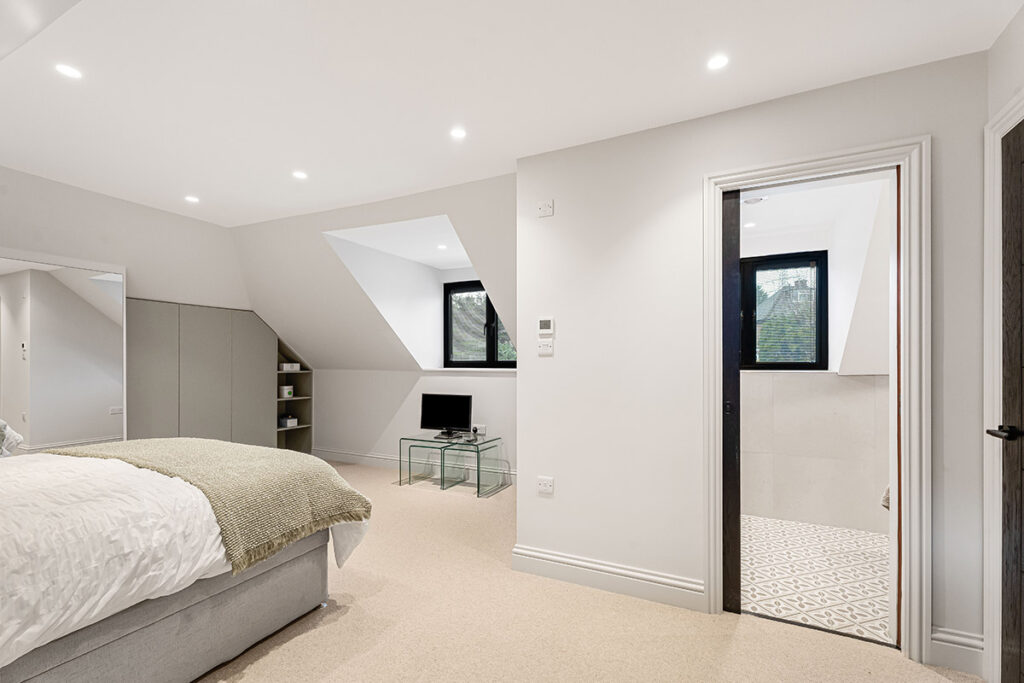 Modern bedroom with a neatly made bed, light carpet, small glass desk with a TV, large wardrobe, and an en-suite bathroom visible through an open door. White walls and ceiling with recessed lighting create a bright atmosphere.
