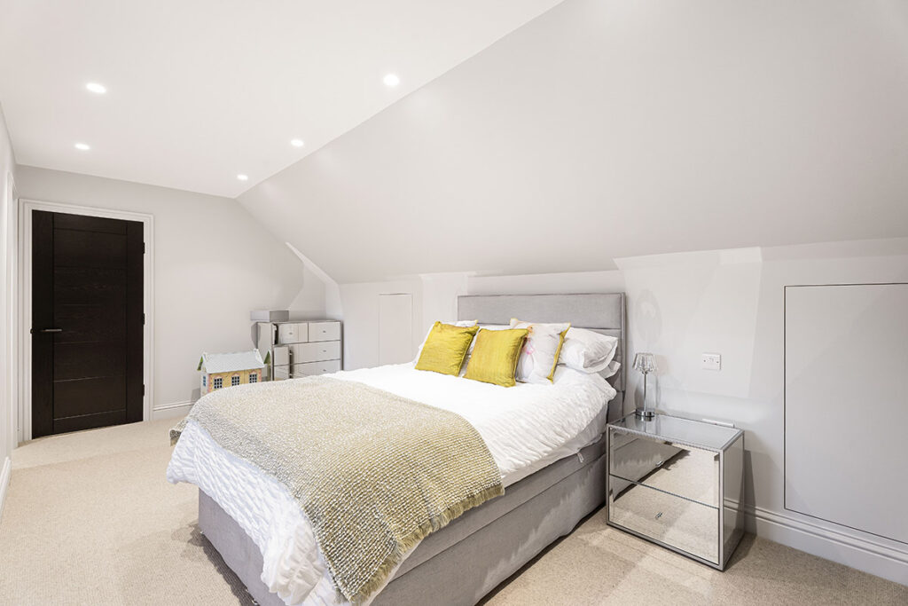 A bright, modern bedroom with a sloped ceiling, white walls, a grey bed with white linens and green accent pillows, mirrored nightstands, and a small dresser. Recessed lighting illuminates the space.