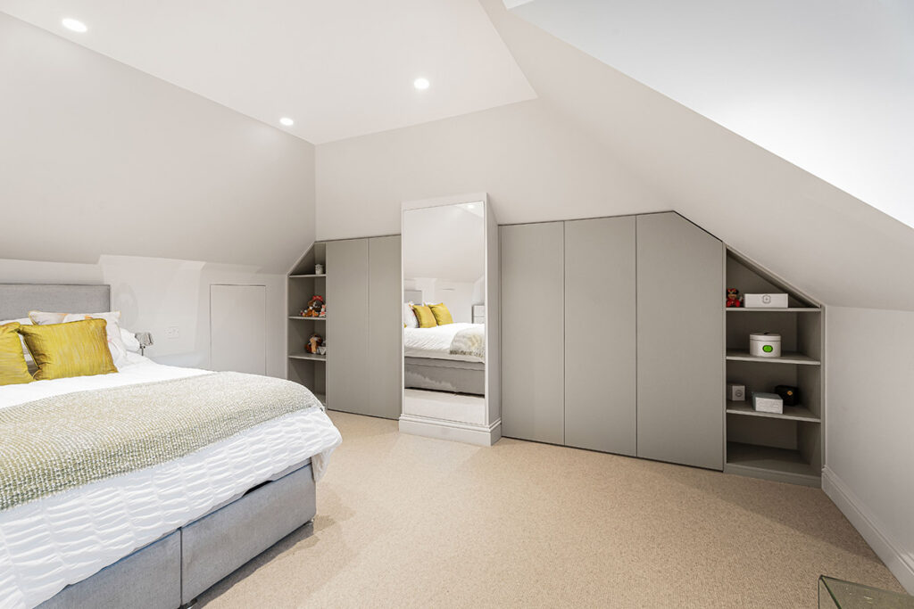 Modern attic bedroom with sloped ceilings, built-in grey wardrobes, open shelving, a tall mirror, and a neatly made bed with white bedding and yellow accent pillows. The space is bright and minimalist.
