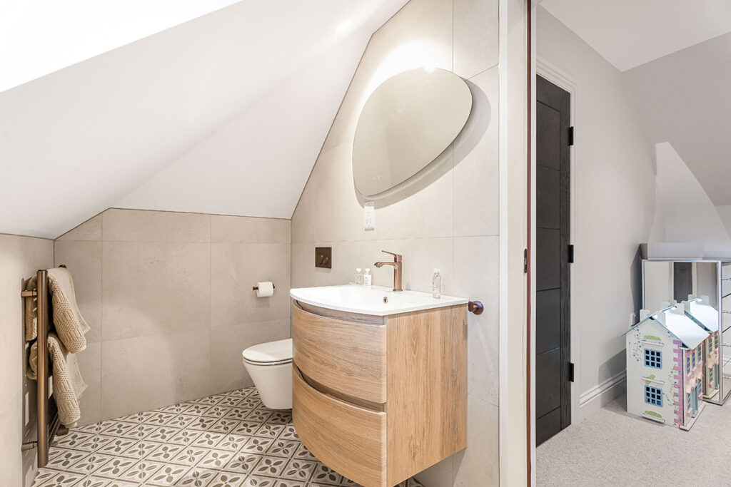 A modern attic bathroom with sloped ceiling, oval mirror, wall-mounted sink cabinet, patterned tile floor, and a towel rack. An adjacent room has a dollhouse and a toy kitchen set.