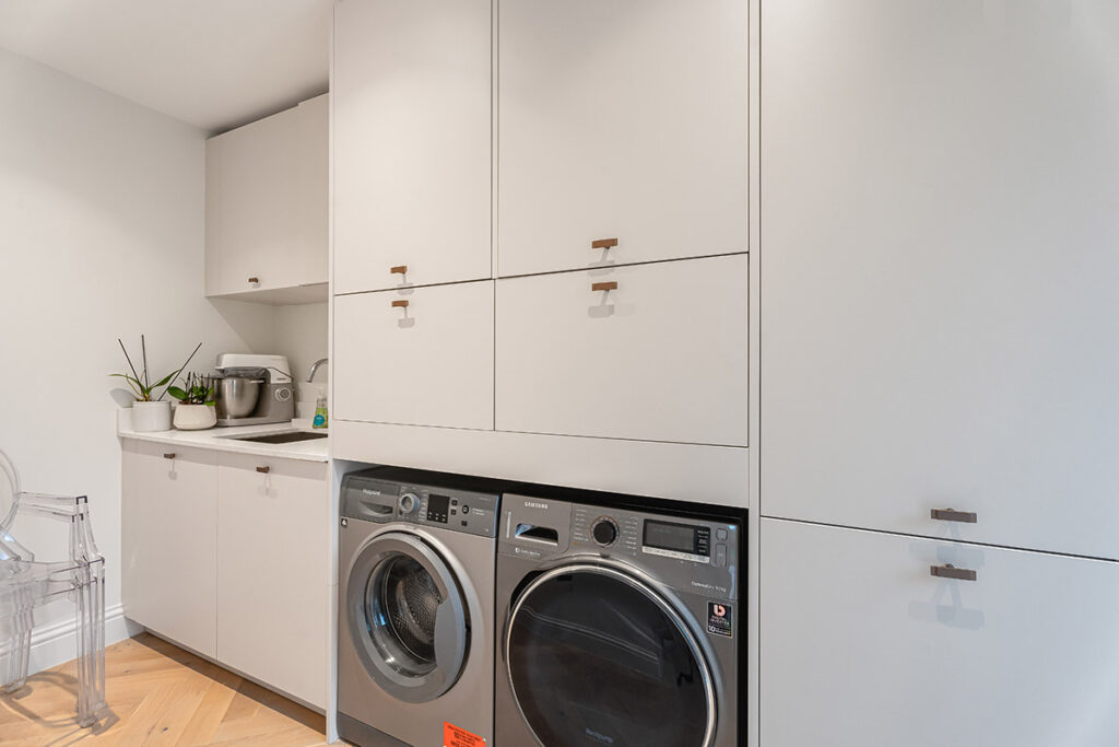 A modern laundry room with white cabinets, a washing machine and dryer, a clear chair, a countertop with a mixer, potted plants, and a sink. The space is clean, bright, and organized.