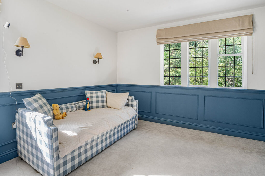 A bright room with a chequered daybed, beige carpet, blue panelled walls, two wall lamps, a window with beige curtains, and soft toys on the bed.