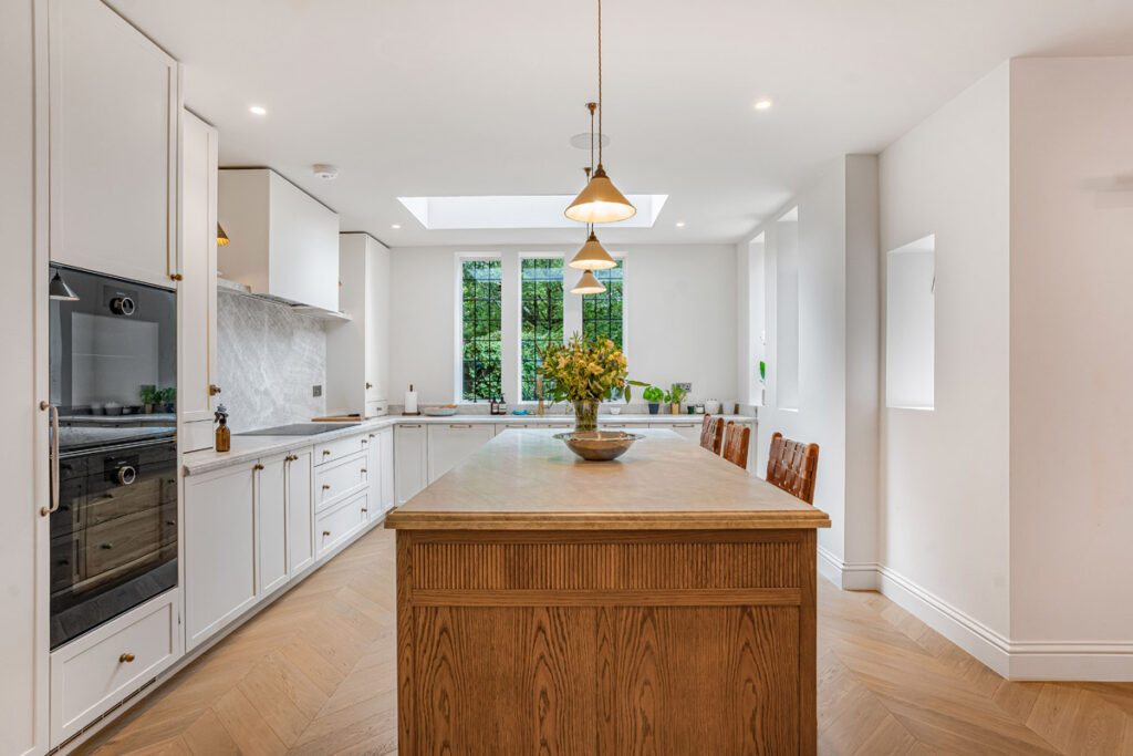 Modern kitchen with white cabinets, built-in oven, marble splashback, and a large wooden island. Pendant lights hang above the island with a vase of yellow flowers. Natural light comes in from windows and a skylight.