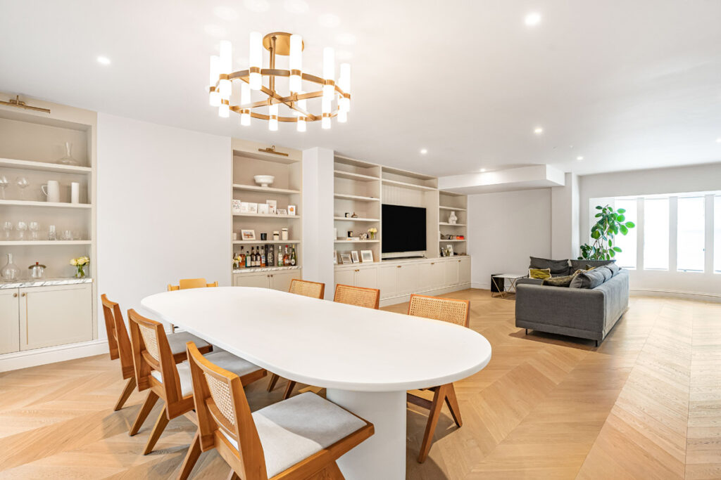 Modern open-plan living and dining area with wooden chairs, white oval table, built-in shelves, a grey corner sofa, large windows, and a gold chandelier. The room is bright with wood-patterned floors.