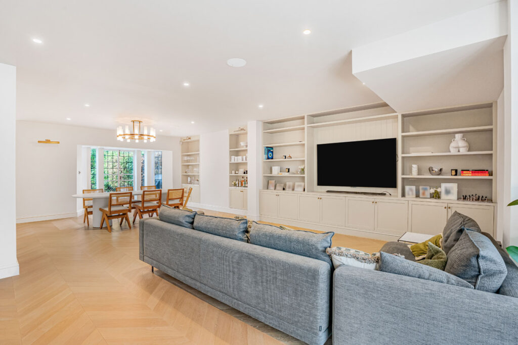 Modern living room with a large grey sectional sofa, built-in shelves, a flat-screen TV, and a dining area with a wooden table and chairs. Light wood flooring and white walls create a bright, open space.