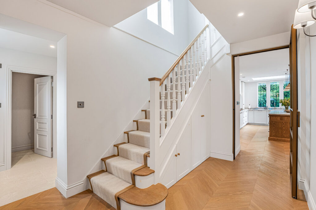 Bright hallway with wooden herringbone floor, white staircase with carpet runner, and open doorway leading to a kitchen with white cupboards and large windows.