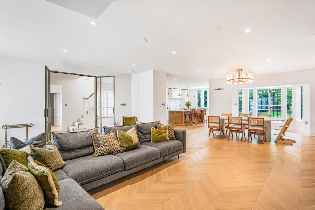 Spacious open-plan lounge with grey sofas, green cushions, wooden flooring, dining area with table and chairs, large windows, and modern kitchen in the background. Bright, airy, and contemporary interior design.