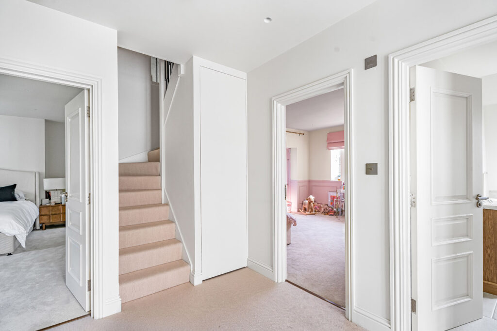 Bright hallway with beige carpet, leading to a bedroom on the left, a staircase in the centre, and a pink playroom on the right. White walls and doors create a clean, modern look.
