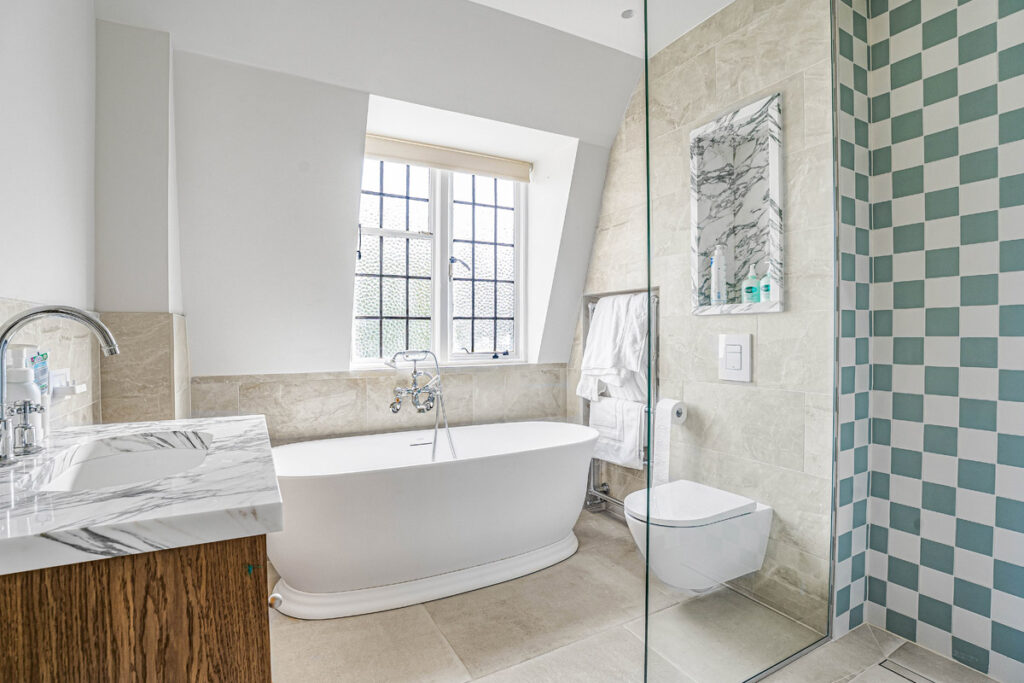 Modern bathroom with a white bath under a window, marble basin, wall-mounted toilet, glass shower area, and blue-grey chequered tiles on one wall. Towels hang on a rail near the bath.