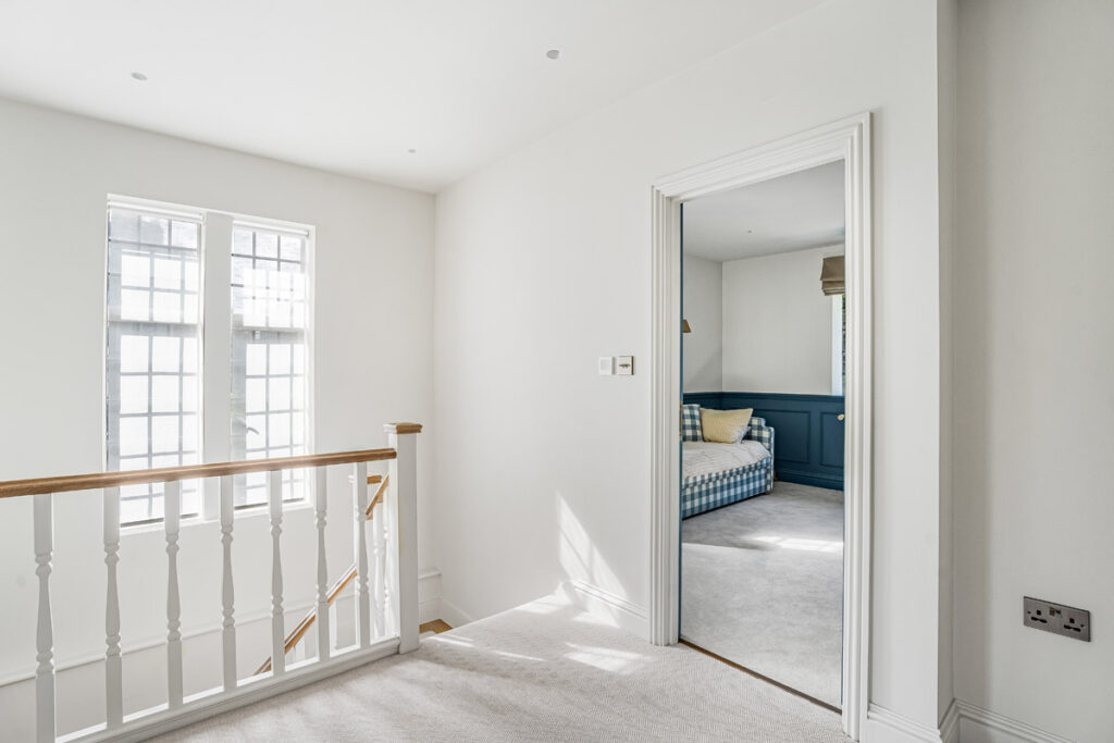Bright hallway with white walls, a carpeted floor, and a staircase with a wooden handrail. An open door reveals a cosy room with blue panelling and a checkered sofa with cushions. Sunlight streams through tall windows.