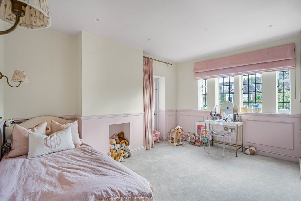 A pastel pink and white children's bedroom features a bed, soft toys, toys, a small table with a mirror, and large windows letting in natural light.