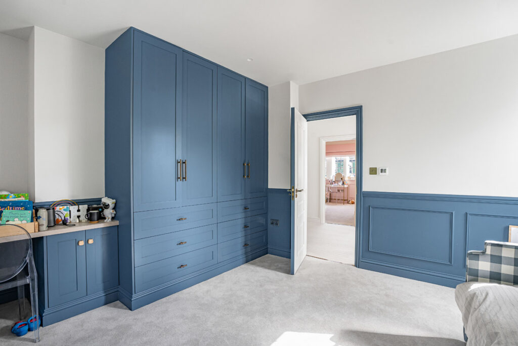 A bedroom with blue fitted wardrobes, blue trim, a desk with toys on the left, a grey carpet, and an open door leading to a sunlit room with pink curtains.