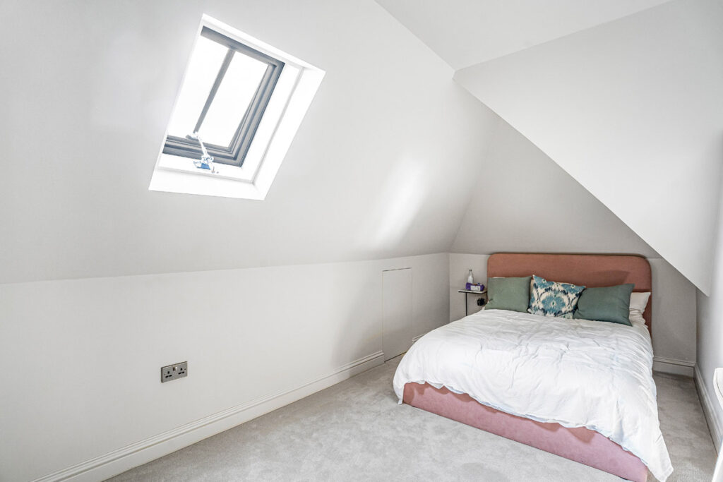 A small attic bedroom with a sloped ceiling, skylight window, and a bed with a blush pink headboard, white bedding, and decorative cushions. A small bedside table and neutral carpet complete the simple decor.