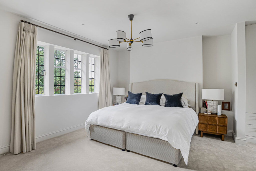 Bright bedroom with large bed, white bedding, dark accent cushions, two bedside tables with lamps, beige carpet, cream curtains, and a modern chandelier. Tall windows let in natural light and offer a view of greenery outside.