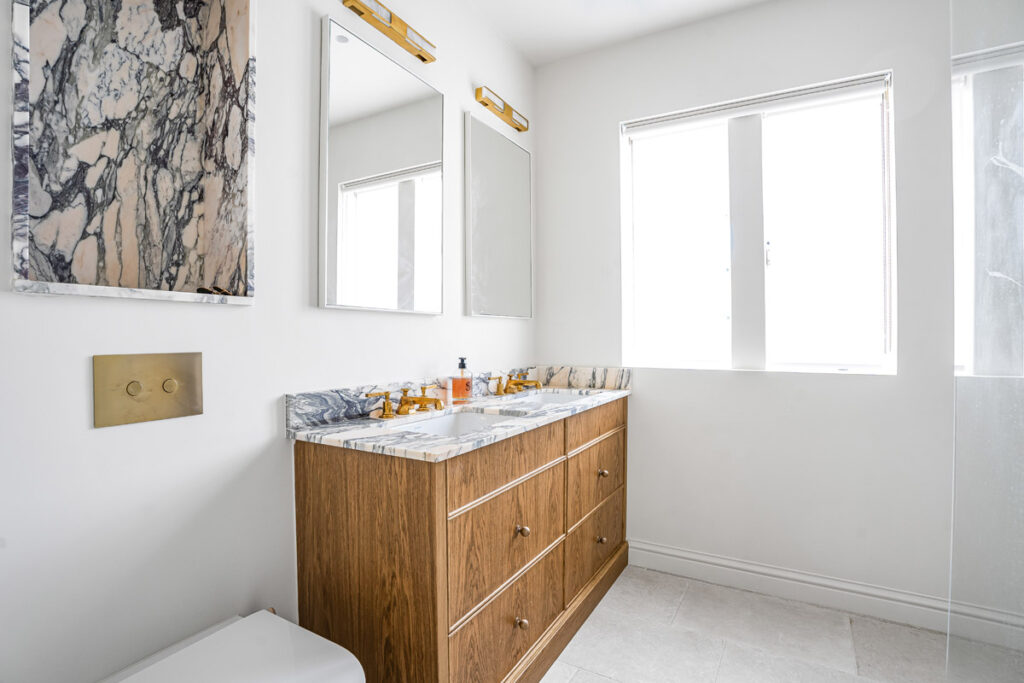 A modern bathroom with a marble countertop, wooden vanity, two mirrors, gold light fittings, a window, and white walls. Marble accents are above the toilet and counter.