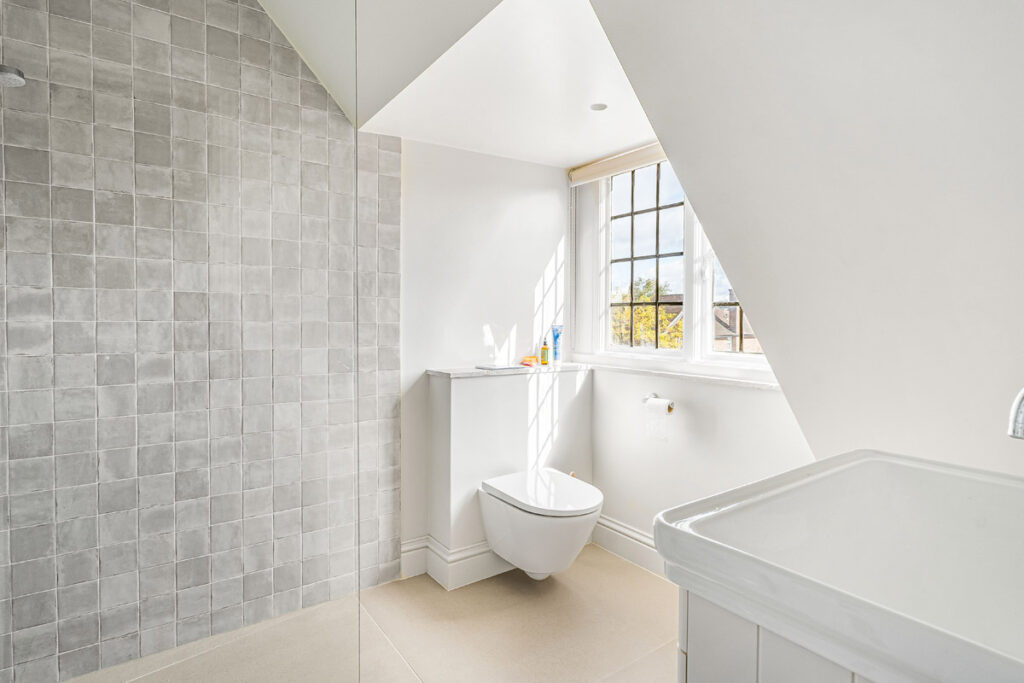 Minimalist bathroom with light grey tiled wall, floating toilet, white basin, and slanted ceiling. Sunlight streams through a window above the toilet, illuminating the bright, clean space. Toiletries sit on the windowsill.