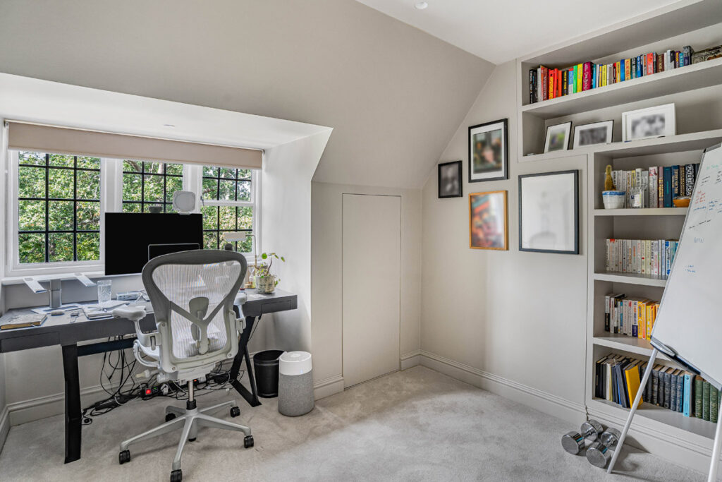 Modern home office with a desk, computer, and office chair by a window. Bookcases, a whiteboard, framed pictures, and potted plants are visible. Room is tidy and well-lit by natural light.