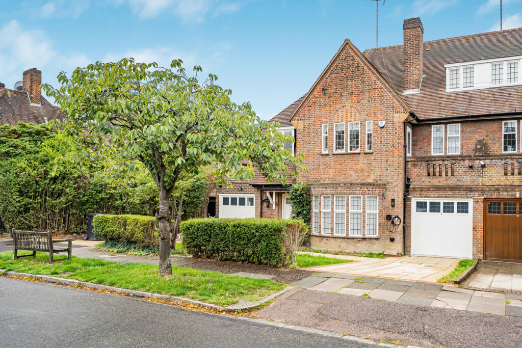 A brick terraced house with a garage, large windows, and a tree in the front garden, situated on a quiet street with a pavement and a wooden bench.