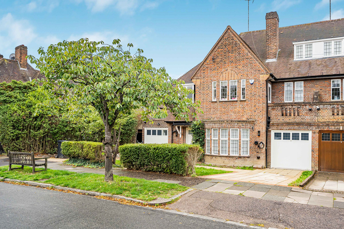 A brick terraced house with a garage, large windows, and a tree in the front garden, situated on a quiet street with a pavement and a wooden bench.
