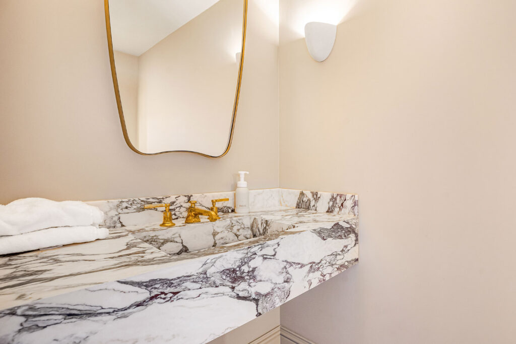 A modern bathroom with a marble sink, gold tap, towel, and a soap dispenser. A curved mirror hangs above and a wall sconce light is mounted on the beige wall.