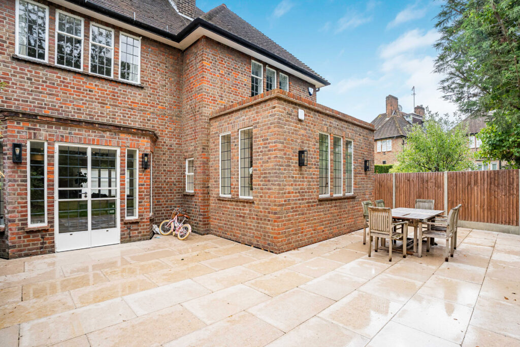 A brick house with large windows opens to a spacious patio. There’s a wooden dining set on the tiled ground and two children’s bicycles near the door. A wooden fence and greenery are in the background.