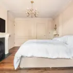 A bedroom featuring a bed with white bedlinen, a fireplace with a wall-mounted television above it, and a gold chandelier.