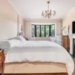 A bedroom with a large bed, a wooden chest of drawers, a fireplace with a mounted television, and a bay window.
