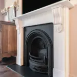 An ornate white fireplace mantel framing a decorative black cast-iron arched firebox, with a wooden chest of drawers visible to the left.
