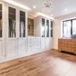 A dressing room with white floor-to-ceiling glass-fronted wardrobes, a wooden chest of drawers beneath a window, and a crystal chandelier.