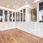 A walk-in wardrobe with white cabinetry, glass-fronted hanging sections, open shelving for accessories, a built-in chest of drawers, and a crystal chandelier.